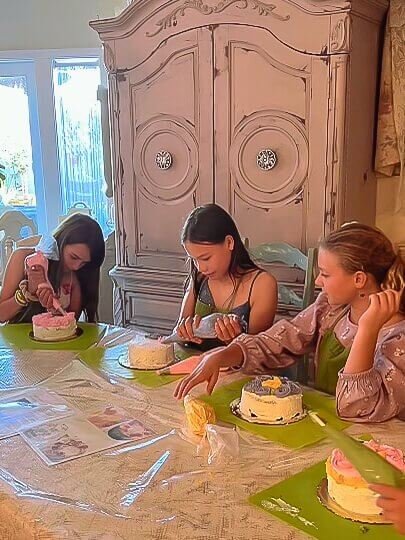 Three girls sit on a table decorating their cake. One has finished, while two are in progress.